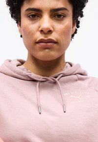 Κοντινό πλάνο ενός ατόμου με κοντά σγουρά μαλλιά που φοράει μια ανοιχτό ροζ φούτερ Puma Athletics με κορδόνια, μπροστά από φόντο σε απλό χρώμα.