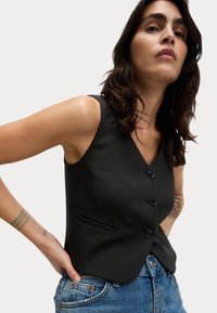 Gilet nero senza maniche con scollo a V, dotato di due bottoni, una tasca frontale e una vestibilità sartoriale, abbinato a jeans di denim blu.