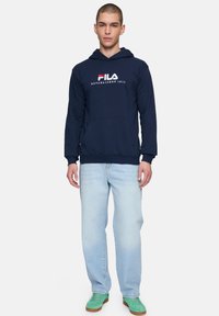 Sudadera azul marino con bolsillo frontal y logo de Fila en blanco, combinada con jeans azul claro y zapatos verdes. Un look casual con capas.