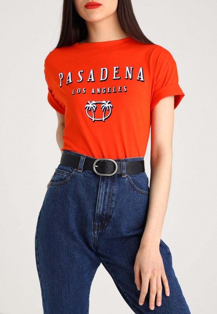 Femme portant un t-shirt orange vif à manches retroussées avec le texte "Pasadena Los Angeles" et un motif de palmier, associé à un jean bleu taille haute et une ceinture noire.
