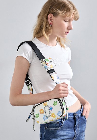 Giovane donna in t-shirt bianca e jeans che tiene una borsa a tracolla bianca con motivi floreali colorati e una spessa tracolla nera.