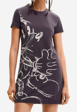 Frau trägt ein kurzärmeliges, dunkles Kleid mit einer großen, weißen, skizzenhaften Grafik von der behandschuhten Hand und dem Gesicht einer Comicfigur.