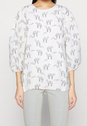 Weiße Bluse mit schwarzen Kamel- und wellenförmigen Linienmustern, aufgebauschte Dreiviertelärmel mit Bindedetails, getragen zu hellgrauen Hosen.