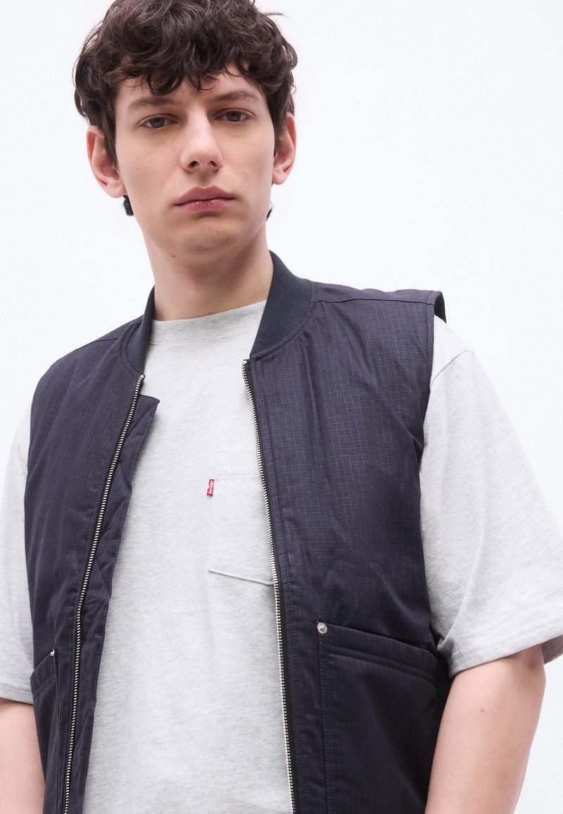 Giovane uomo con capelli scuri ricci, indossa una maglietta grigio chiaro e un gilet senza maniche blu scuro con zip, con sfondo bianco neutro.