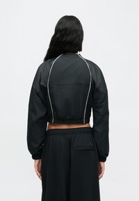 Κοντό μαύρο τζάκετ με ελαστική μέση, διακοσμημένο με λευκή απόληξη κατά μήκος των ραφών και φουσκωτά μανίκια. Συνδυασμένο με φαρδιά μαύρα παντελόνια.