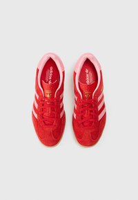 Sarkanas zamšādas sporta kurpes ar rozā akcentiem, trīs baltām svītrām, apaļu purngalu un teksturētu gumijas zolīti. Zelta logotipa detaļas uz mēles.