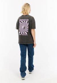 Donkergrijze oversized t-shirt met een lichtpaarse spiraalafbeelding op de achterkant, gedragen met blauwe denim jeans en sneakers. De tekst zegt "SUNNY VIBES FOREVER."