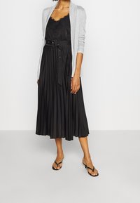 Czarna plisowana sukienka midi z paskiem w talii, zestawiona z jasnoszarym kardiganem. Dopełnieniem stylizacji są sandały i top z koronkowymi wykończeniami.