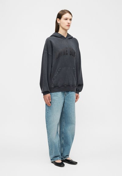 Femme aux longs cheveux bruns portant un sweat à capuche noir ample avec poche avant, un jean large bleu clair, et des ballerines noires à bouts pointus, debout.