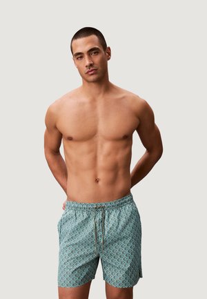 Homme torse nu aux cheveux courts portant un short de bain bleu clair à motifs, debout les mains dans le dos devant un fond uni.