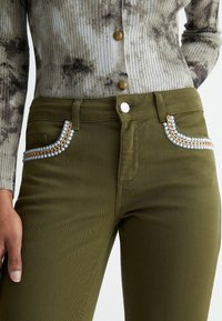 Jeans ajustados color verde oliva con bolsillos decorativos con cadena dorada y detalles de cuentas azul claro, combinados con una camiseta acanalada con estampado.