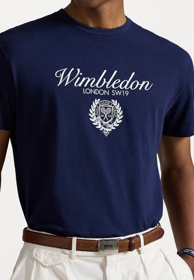 Tricou din bumbac de culoare bleumarin, cu text alb „Wimbledon LONDON SW19” și un logo cu frunze de dafin și rachete de tenis. Mâneci scurte.