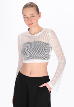 Junge Frau mit zurückgebundenen blonden Haaren trägt ein weißes, langärmliges, bauchfreies Netzoberteil über einem schwarzen Bandeau, kombiniert mit schwarzen Hosen.