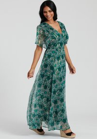 Robe maxi en tissu turquoise transparent avec un motif floral. Présente des manches courtes évasées, un décolleté en V et une taille cintrée. Portée avec des sandales compensées.