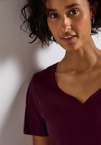 Burgundská trička s v-výstřihem, krátkým rukávem, hladkou texturou a přiléhavým střihem, na kterém je vidět osoba s kudrnatými vlasy na neutrálním pozadí.