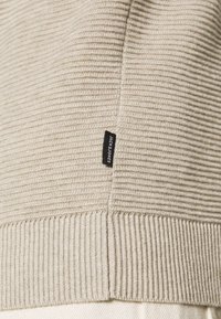 Détail d'un pull beige à côtes avec une couture visible et une étiquette en tissu noir sur le côté, comportant du texte. La texture semble douce et texturée.
