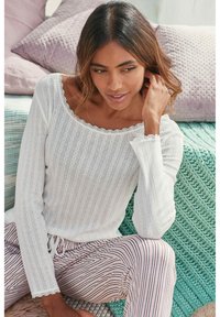 Maglia bianca a maniche lunghe con scollatura ondulata e righe verticali testurizzate, abbinata a pantaloni a righe rosa e bianchi. Aspetto morbido e informale.