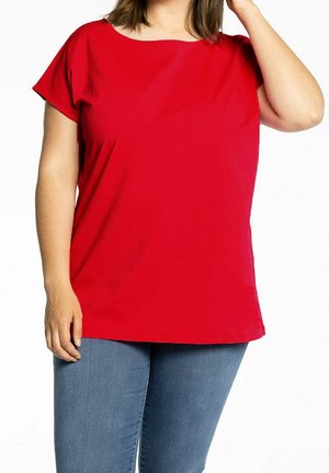 Frau trägt ein schlichtes rotes Kurzarmshirt und blaue Jeans, mit einer Hand an der Seite und der anderen nahe am Kopf.
