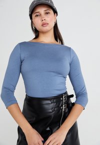 Μακρυμάνικο, ανοιχτό μπλε μπλουζάκι φτιαγμένο από μαλακό ύφασμα, συνδυασμένο με ψηλόμεση φούστα από ψεύτικο δέρμα σε μαύρο χρώμα, που διαθέτει διπλές αγκράφες.