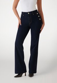 Hoch taillierte dunkelblaue Jeans aus Denim mit Seitenschbuttons, weitem Bein und Kontrastnähten. Getragen mit schwarzen spitzen Stilettos.
