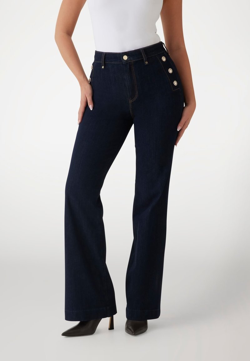 Hoch taillierte dunkelblaue Jeans aus Denim mit Seitenschbuttons, weitem Bein und Kontrastnähten. Getragen mit schwarzen spitzen Stilettos.