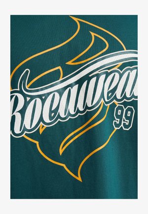 Roheline kangast T-särk, millel on valge ja kollane "Rocawear" logo koos tulekujundusega ning all numbriga 99. Pehme tekstuur, juhuslik stiil.