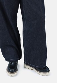 Pantalones de mezclilla oscuros de pierna ancha con costuras blancas, combinados con zapatos de cuero negros que tienen una suela gruesa beige.