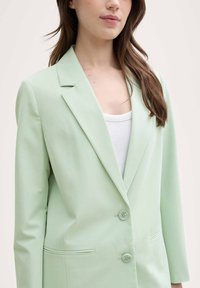 Hellgrüner Blazer mit strukturiertem Design, zwei Knöpfen und Reverskragen. Das Material wirkt glatt mit minimaler Textur.