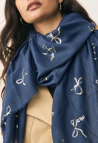 Sciarpa blu navy con tessitura strutturata e motivi di fiocchi dorati, indossata intorno al collo, con bordi con frange e tessuto morbido.