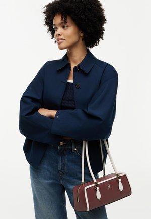 Femme aux cheveux bouclés portant une veste bleu marine et un jean, tenant à son côté un sac à main rectangulaire bordeaux avec des anses blanches.