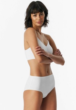 Frau mit dunklem Haar, die ein weißes Bralette und hoch taillierte Slips trägt, steht mit verschränkten Armen vor einem weißen Hintergrund.