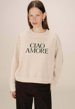 Sweat-shirt beige avec "CIAO AMORE" imprimé en vert foncé, présentant une coupe décontractée et un col et des poignets côtelés, associé à un pantalon sombre.