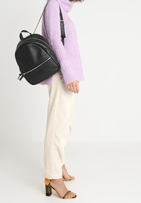 Kvinna i lila tröja, krämfärgade byxor och gult randiga klackar som bär en svart läderryggsäck över ena axeln.