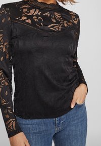 Sort langærmet top med blomsterbroderi, høj halsudskæring, rynkede detaljer og tekstureret stof. Sammen med blå denimjeans.