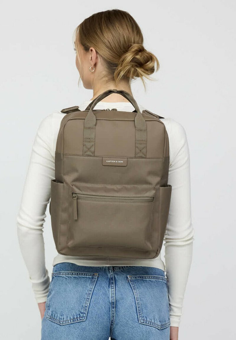 Khaki-Rucksack mit doppelten Obergriffen, gepolsterten Schulterriemen, einer vorderen Reißverschlusstasche und Seitentaschen. Glattes Material mit einer strukturierten Form.