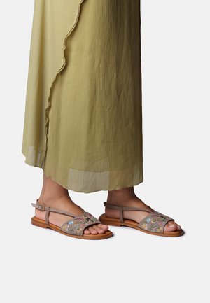 Noge u bež sandalama s cvjetnim vezom i trakama oko gležnja, djelomično prekrivene dugom, svijetlozelenom lepršavom haljinom.