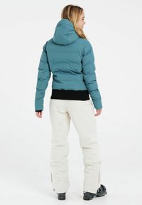 Türkise Pufferjacke mit Kapuze, cropped Design und geripptem schwarzen Bund. Getragen mit weißen Hosen und schwarzen Skischuhen.