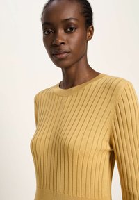 Nahaufnahme einer Frau, die einen senfgelben, gerippten Strickpullover mit Rundhalsausschnitt trägt, vor einem schlichten hellen Hintergrund.