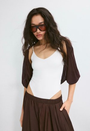 Kvinde med langt mørkt hår iført store brune solbriller, hvid ærmeløs bodystocking, løs brun nederdel og en brun shrug med pufærmer.