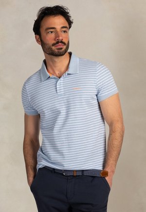 Man met baard, gekleed in een lichtblauw-wit gestreept poloshirt en een donkere broek, met handen in de zakken, kijkt iets zijwaarts.