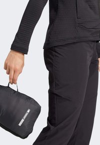 Haut noir à manches longues avec un motif texturé et des manches ajustées, associé à un pantalon noir léger. La personne tient une veste compacte et pliée.