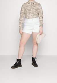 Beige skjorta med zebramönster, vita denimshorts och svarta snörstövlar. Vit kuvertväska hålls i höger hand.