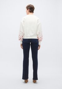 Λευκό φούτερ με κόκκινα σχέδια καρδιών στα μανίκια, συνδυασμένο με σκούρο μπλε τζιν με καμπάνα. Απλός σχεδιασμός, απαλό ύφασμα, άνετη εφαρμογή.