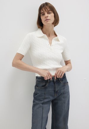 Woman with short brown hair wearing white short-sleeve cable knit polo shirt and high-waisted loose blue jeans, adjusting jeans waistband.