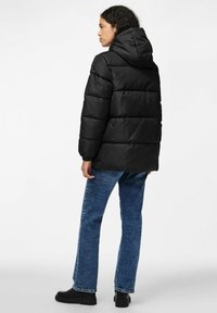 Svart puffjacka med huva, som har ett quiltat mönster och elastiska mudd. Kombinerad med blå denimjeans och svarta anklarstövlar.