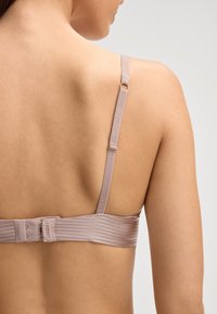 Schiena nuda di una donna che indossa un reggiseno beige con spalline regolabili e chiusura a gancio su uno sfondo chiaro e semplice.