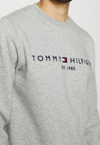 Sudadera gris con cuello redondo, con el logo "TOMMY HILFIGER" en azul marino, rojo y blanco. Tejido suave con un ajuste clásico.
