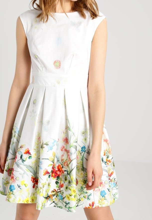 Robe blanche sans manches avec un imprimé floral coloré le long de la jupe, portée par une personne debout sur un fond uni.
