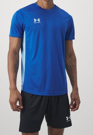Mand iført blå og lyseblå Under Armour kortærmet skjorte og sorte Under Armour shorts stående mod en ensfarvet baggrund.