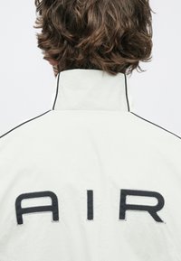 Λευκό μπουφάν με ψηλό γιακά, διακοσμημένο με μαύρα ραφές και μεγάλα γράμματα που σχηματίζουν τη λέξη "AIR" στην πλάτη. Λείο, ελαφρύ υλικό.